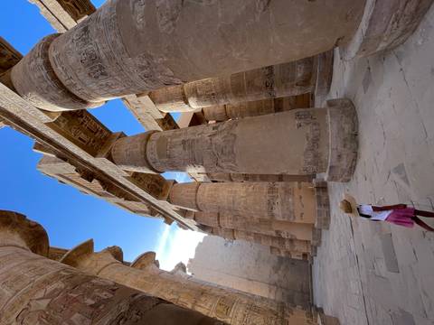 Young girl in a hat walks beneath towering sandstone columns bathed in afternoon light inside Karnak Temple.