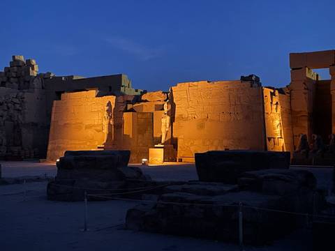 Nighttime illumination of ancient sandstone walls and statues within Karnak Temple complex against a dark blue sky.