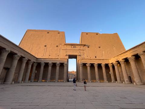 Golden evening light bathes the courtyard of Edfu Temple as two tourists stand beneath monumental walls carved with deities.