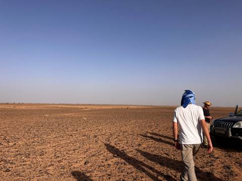 Traveler in white shirt with blue headscarf walks toward companions across a vast barren landscape.