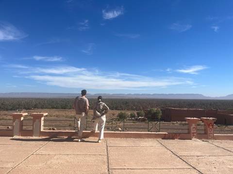 Two travelers pause at a scenic overlook with expansive palm groves and distant mountains beneath a blue sky.