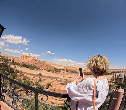 Woman taking a photo of the mud-brick fortified village stretching across a desert valley under a bright blue sky.