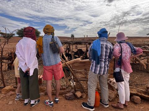 Travelers wearing head scarves observe goats in a rustic pen in a dry desert setting under a partly cloudy sky.