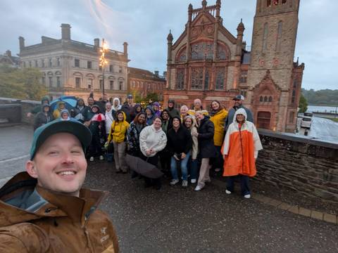 Rain-jacketed group gathers for a selfie on a stone bridge with the ornate red-brick Guildhall in the background.