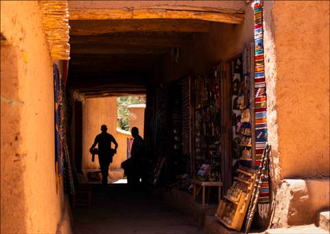 Silhouetted figures walk through a shadowy adobe corridor lined with colorful crafts and sunlight at the far end.