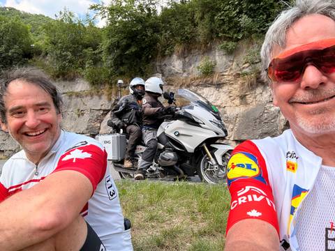 Cyclists chatting roadside as motorcycle camera crew passes in background