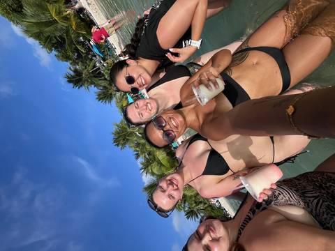 Four women in swimsuits holding tropical drinks while standing in shallow beach water