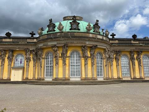 Front view of the yellow Baroque Sanssouci Palace with its green dome under a partly cloudy sky.