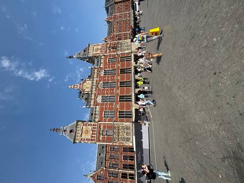 Visitors gather on the square in front of Amsterdam Centraal Station on a bright blue-sky day.