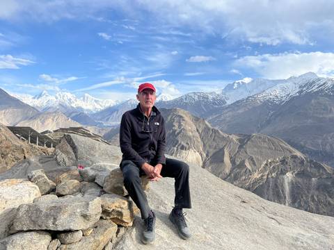 Traveller seated on rocks overlooking vast mountain ranges under a bright sky.