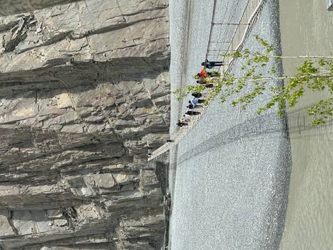 Travellers carefully cross a long cable bridge suspended beside towering rock cliffs.