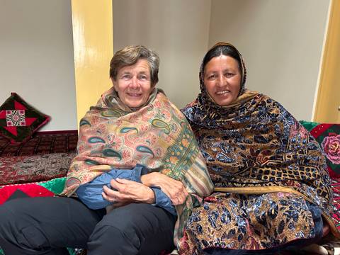 Two women wrapped in patterned shawls smile while seated on vibrant cushions.