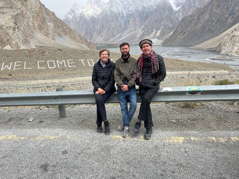 Three travellers rest on a guardrail with a valley view and rock text reading "WELCOME TO" behind.