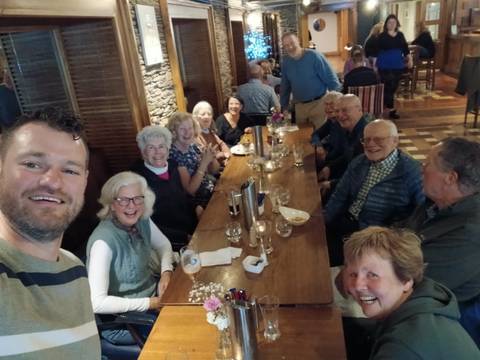 Smiling tour group gathered around a long wooden pub table with drinks and cozy décor.