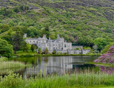Stately castle reflected in a serene lake surrounded by lush green hills and blooming rhododendrons.