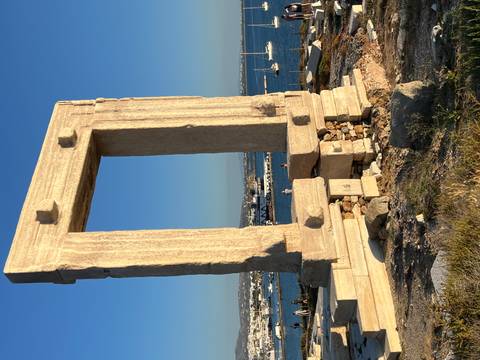 The ancient marble Portara gateway stands on a promontory overlooking the Aegean Sea with sailboats below.
