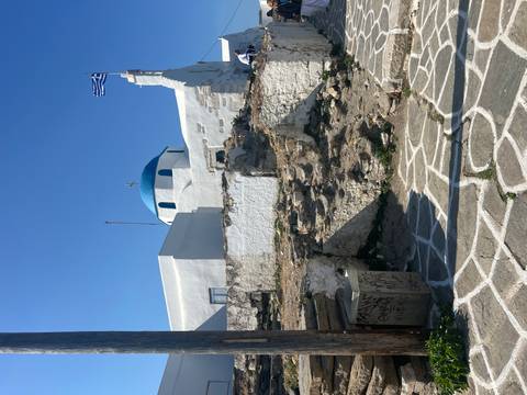 Blue-domed whitewashed church topped with a Greek flag against a clear sky.