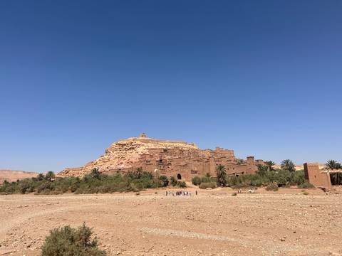 Earthen kasbah of Aït Benhaddou rises above a dry riverbed and palm trees under clear blue sky.
