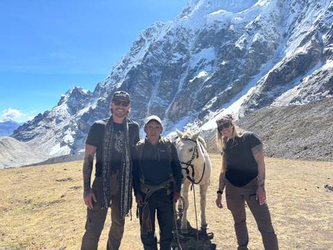 Travellers and a local horse handler posing with a white horse beneath rugged Andean cliffs