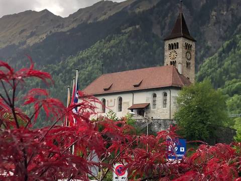 Small alpine church with clock tower set against steep green mountains and red foreground foliage.