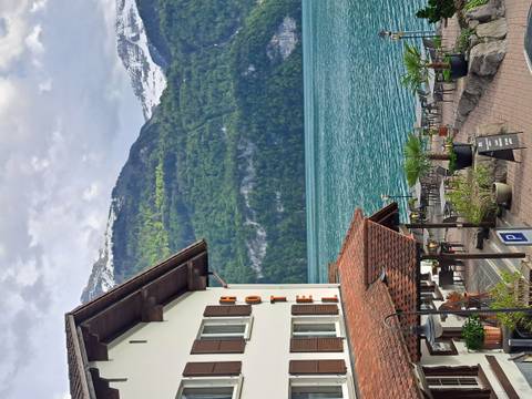 Lakefront alpine hotel with outdoor terrace overlooking turquoise water and snow-capped peaks under a lightly clouded sky.