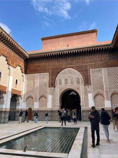 Intricately carved entrance and tiled walls of Ben Youssef Madrasa courtyard with visitors milling about.
