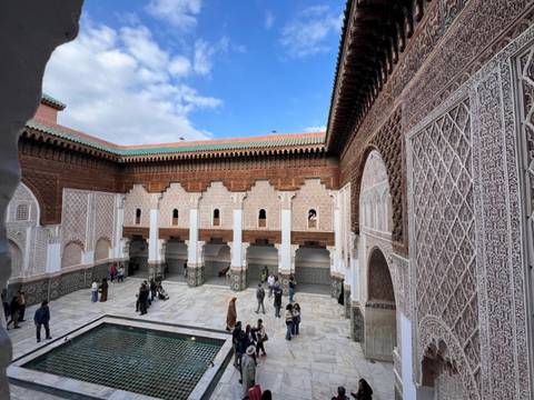 Angled view across the ornate courtyard of Ben Youssef Madrasa with tiled pool, carved arches and visiting tourists.