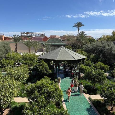 Lush courtyard garden with hexagonal gazebo shaded by orange trees, visitors strolling on green tile paths.