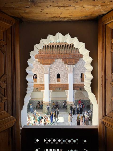 View through a sculpted keyhole window framing the courtyard and arches of a historic madrasa with visitors below.
