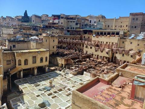 Panoramic view of a traditional tannery with stone vats of dye and leather drying on rooftops, worker visible below.