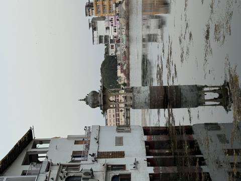 Still lake with historic stone pavilion column reflecting in water beside weathered buildings, Udaipur.
