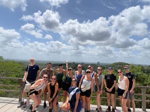 Large tour group poses at a jungle viewpoint overlooking distant Maya temples of Tikal.
