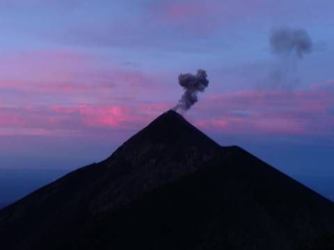 A smoking conical volcano silhouetted against a pink and blue twilight sky.