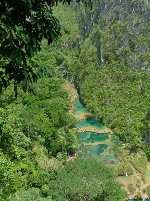 An emerald river with turquoise limestone pools winding through dense tropical jungle seen from above.
