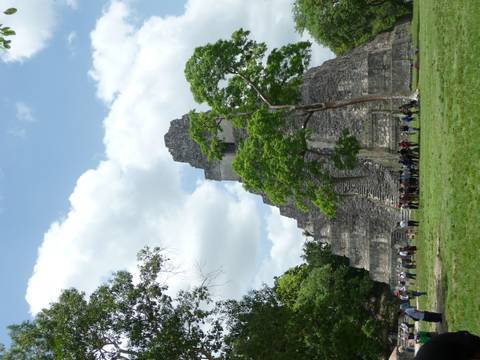 The iconic stepped pyramid of Tikal rises above grassy grounds where tourists gather under a partly cloudy sky.