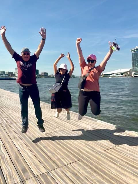 Friends mid-air while jumping from a wooden pier on Amsterdam waterfront with city skyline behind.