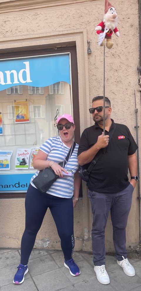 Two travellers pose with playful expressions outside a shop holding umbrellas on a cloudy day.