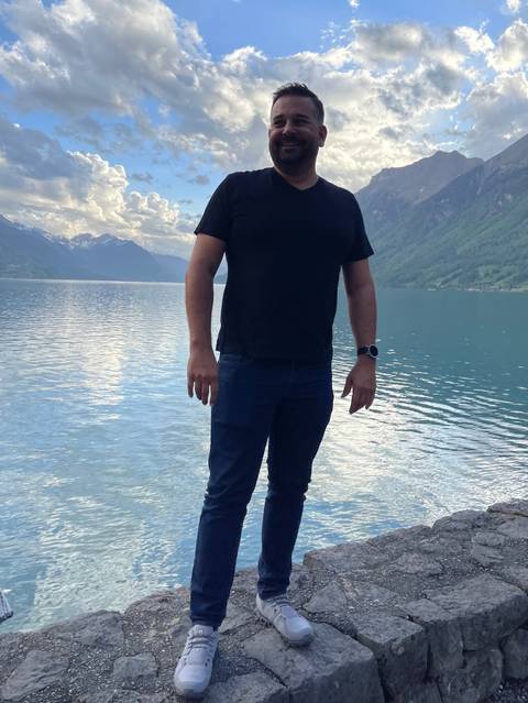 Traveller stands by a tranquil alpine lake with snow-capped peaks in the distance.