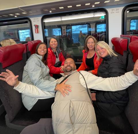 Travellers laugh together while seated on red train seats during an alpine rail journey.