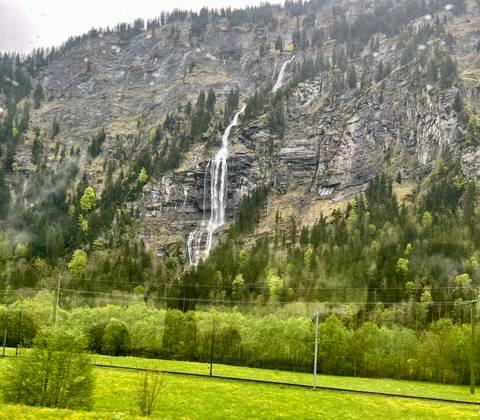 Tall waterfall cascades down misty forested cliffside in the Alps with fresh spring greenery.