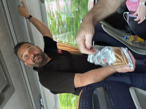 Tour leader enthusiastically offers packaged snacks to passengers on a moving coach bus.