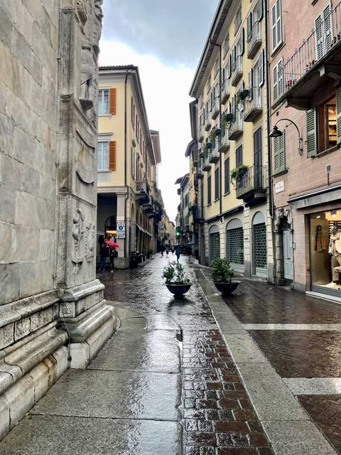 Rain-soaked narrow Italian street lined with pastel buildings and a few pedestrians holding umbrellas
