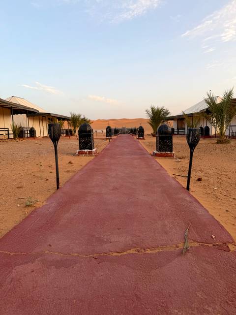 Pathway lined with lantern stands leading to tents in a desert camp with dunes behind.