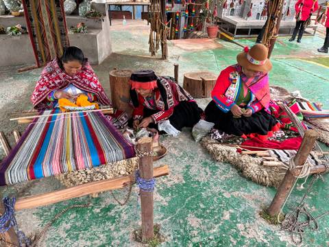 Three Andean women in traditional clothing weave colorful textiles on wooden looms in an open workshop.