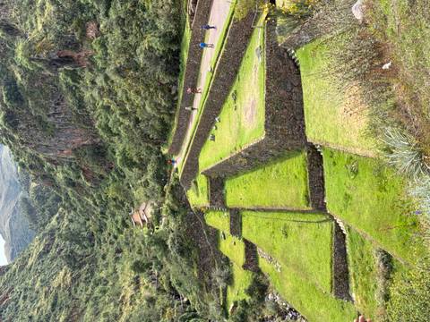 Green terraced fields and stone walls line a winding valley road with hikers in the distance and forested cliffs rising around.
