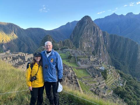 Couple stands together on grassy slope overlooking the iconic ruins and peak of Machu Picchu at dawn light.