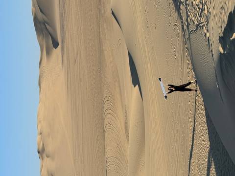 Person holding a sandboard above their head on vast golden dunes under clear sky