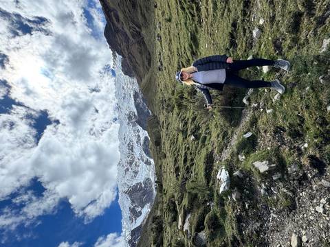 Female trekker with pole on grassy slope beneath snowy Andean peaks and dramatic clouds