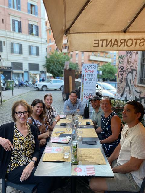 Smiling travellers enjoy drinks around an outdoor table on a cobblestone Roman street lined with scooters and graffiti.