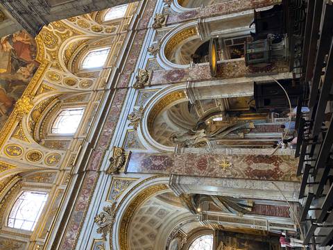 Ornate marble interior of a Renaissance church with gilded arches and soaring painted ceiling; two visitors sit in pews.